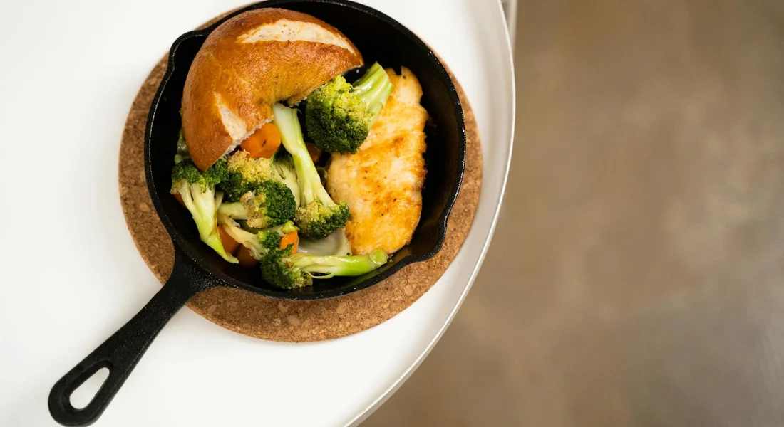 Small cast-iron skillet on a cork trivet containing broccoli florets and a piece of chicken with a bread roll on the side, set on a white plate.