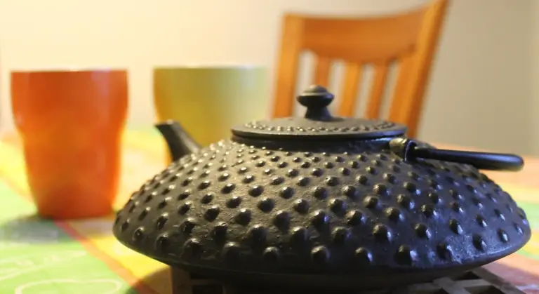 Close-up of a textured cast iron pot lid on a kitchen table with blurred cups and a chair in the background.