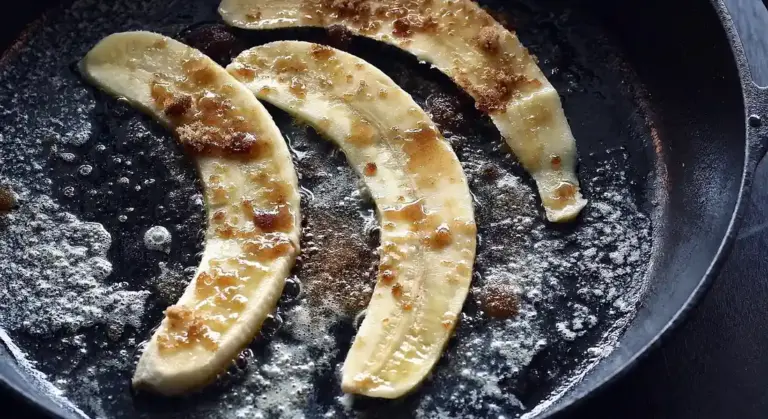Cast-iron skillet with fried banana slices cooking in oil, showing grease and bubbles.