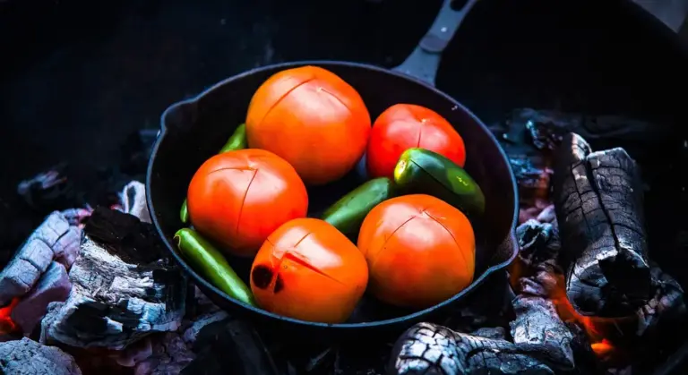Cast iron skillet over glowing embers with red tomatoes and green peppers inside