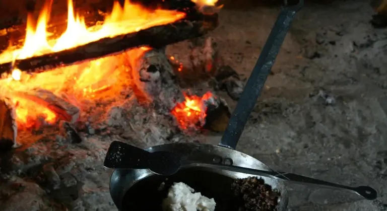 Cast iron pan near a campfire with remnants of food and a long serving spoon
