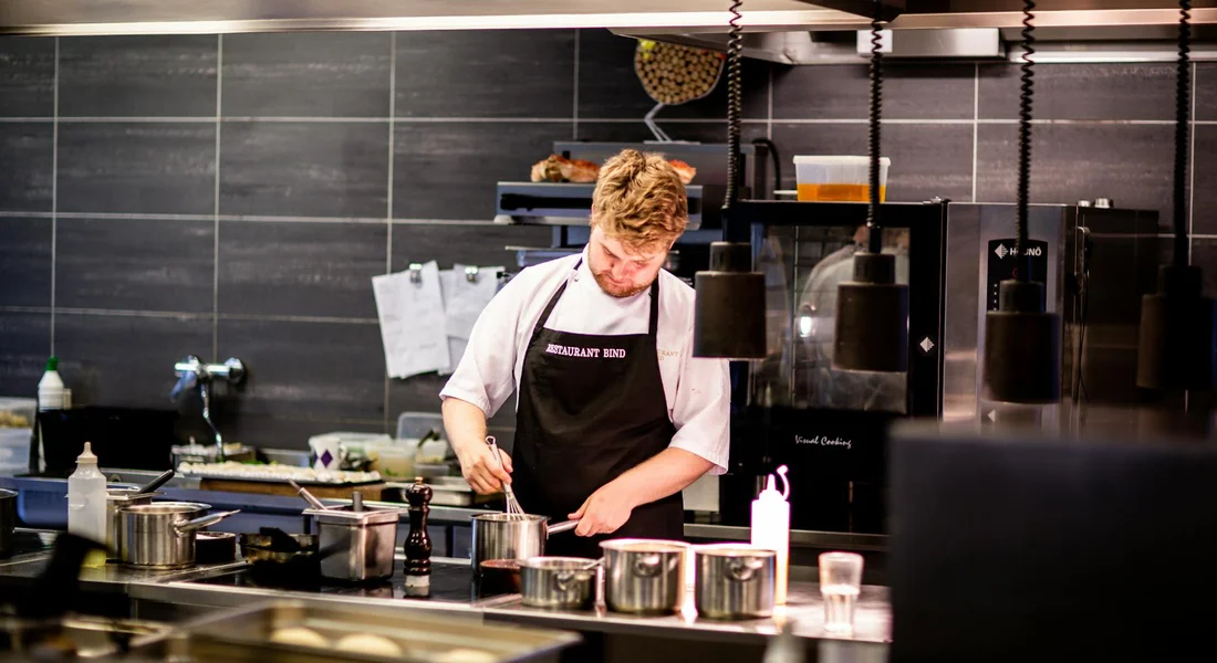 Chef in a professional kitchen at a prep station with pots on the stove, illustrating the post-cleaning phase of drying and preparing cast iron cookware for re-seasoning.