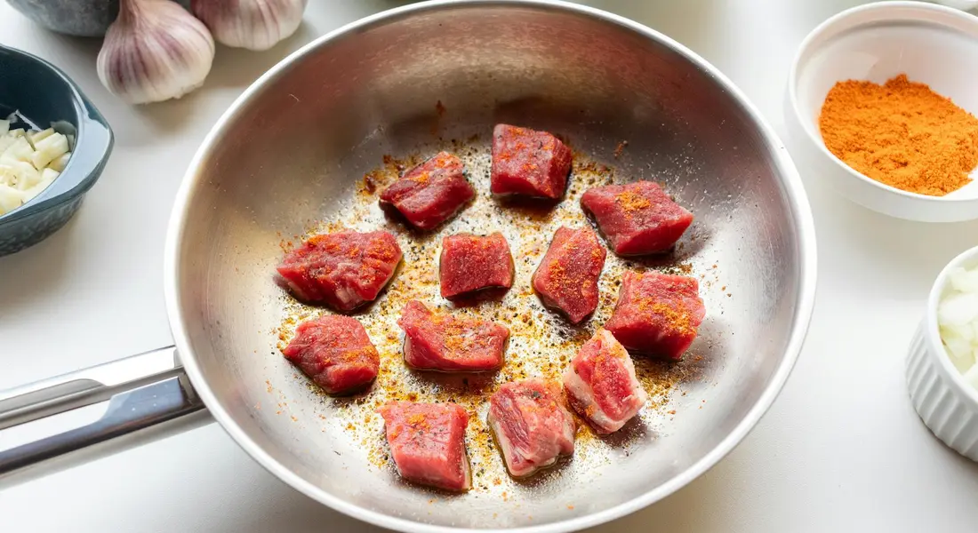 Cast iron skillet with seared pork cubes and seasonings, surrounded by garlic, onions, and bowls of spices.
