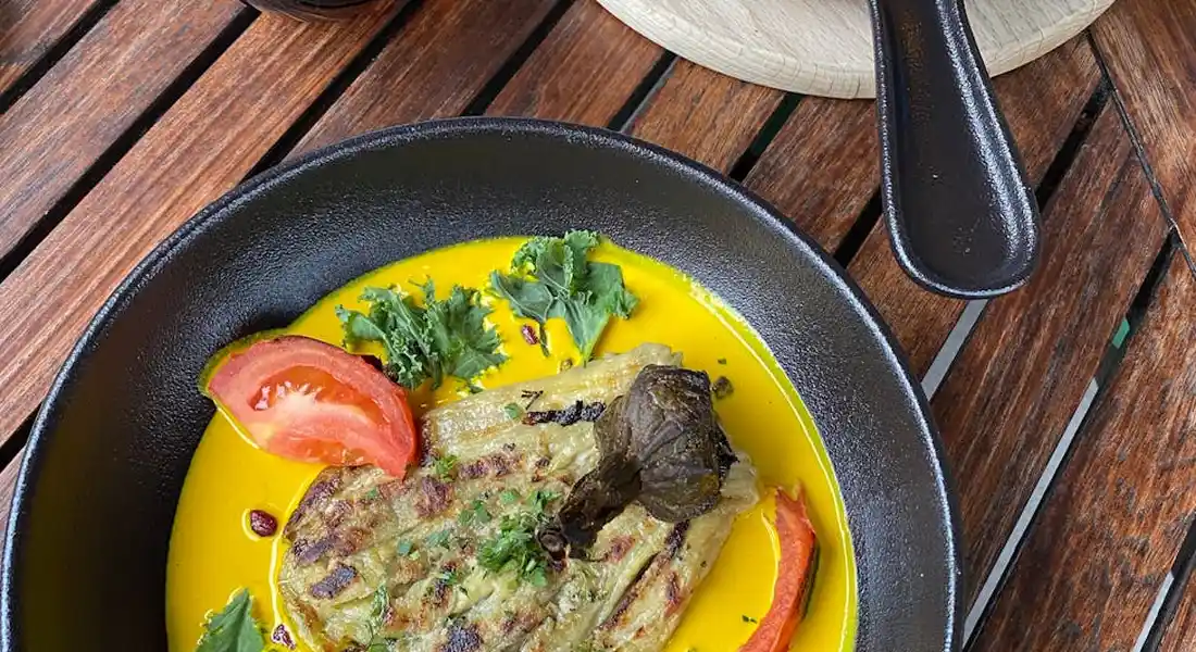 A black cast-iron skillet on a wooden table with a plated dish in bright yellow sauce, garnished with tomato and parsley, and a spatula resting nearby.