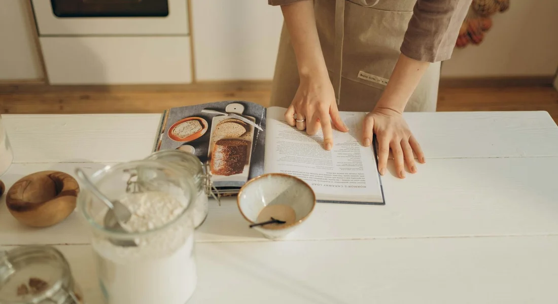 A person stands at a kitchen island, reading a cookbook, with ingredients and cookware laid out, illustrating preparation for cooking with a cast iron Dutch oven.