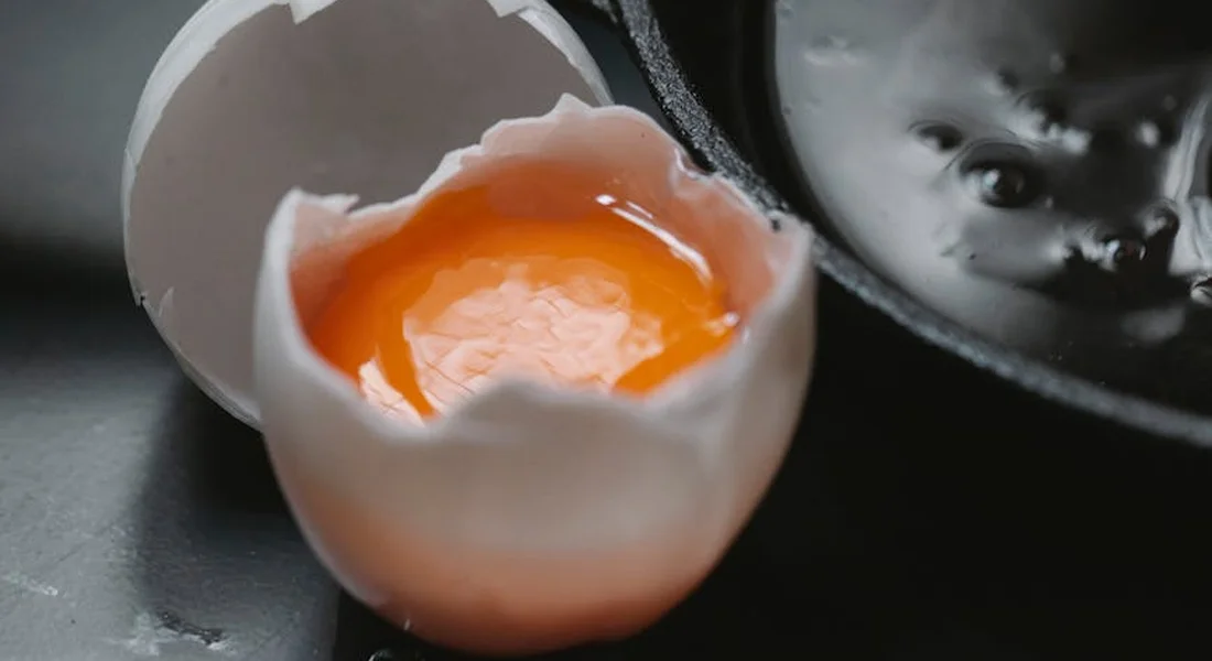 Close-up of a cracked eggshell with a bright orange yolk beside a cast iron skillet, with another skillet visible in the background.