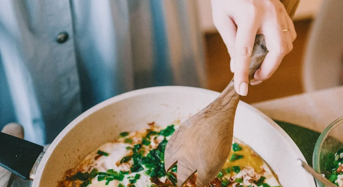 A person stirring a simmering dish in a light-colored cast iron skillet with a wooden spatula, illustrating daily maintenance while cooking.