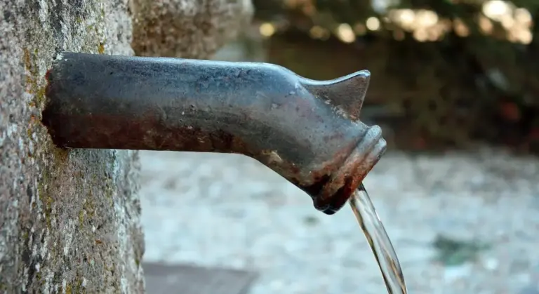 Close-up of a rusty wall-mounted spout with a stream of water dripping from its tip.