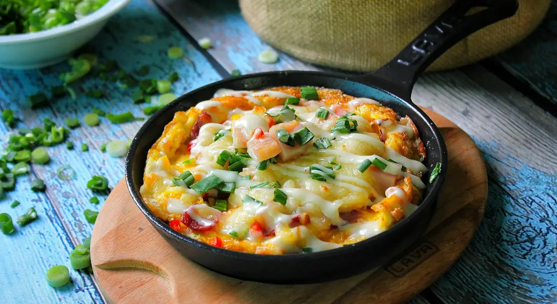 Cast iron skillet with a cheesy egg scramble on a wooden board; herbs and a blue textured surface in the background