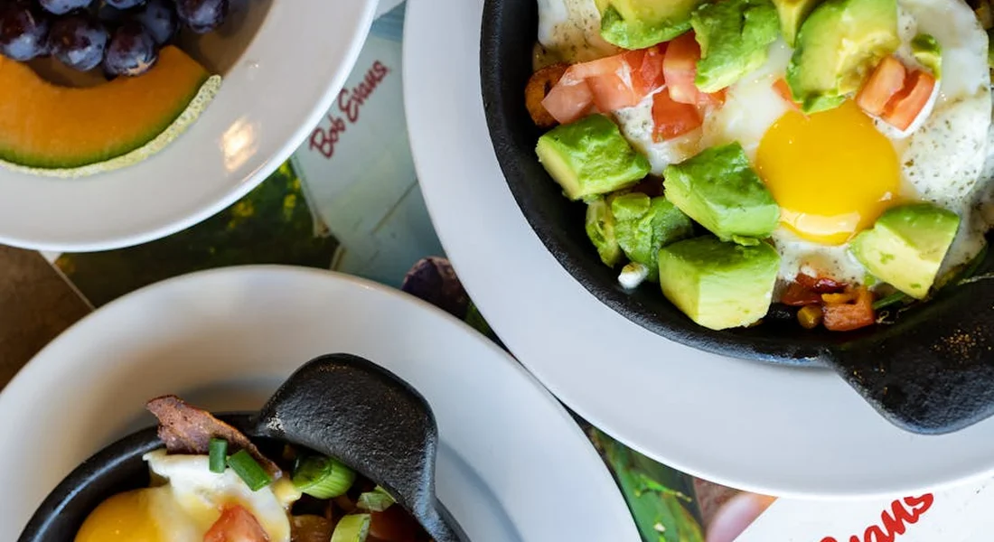 Top-down view of two enameled cast iron skillets on plates, each with a breakfast featuring a sunny-side-up egg, avocado, and tomatoes.