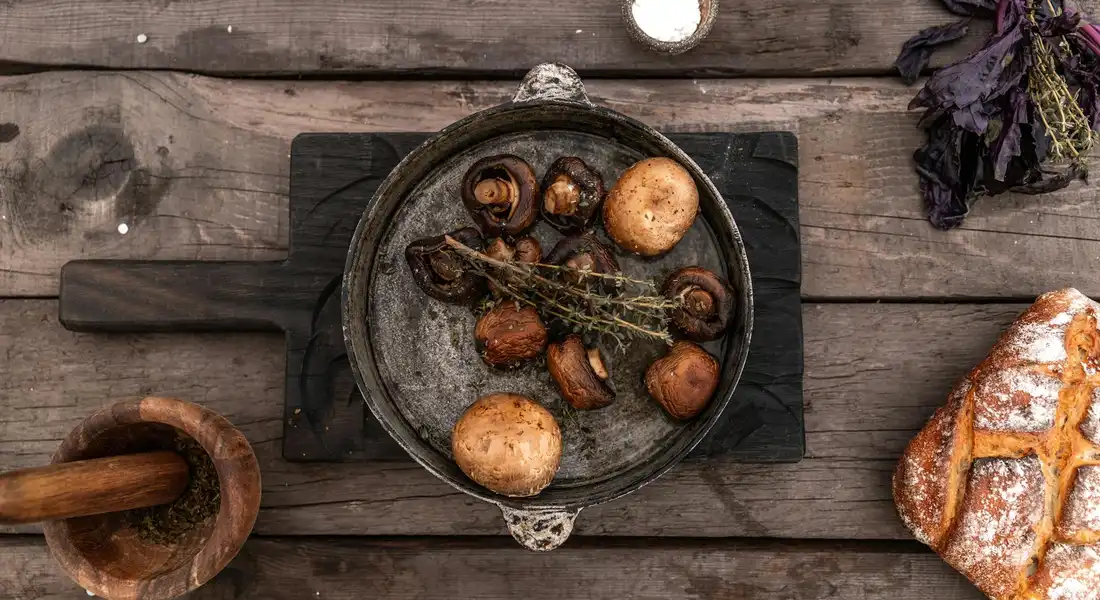 Cast iron skillet with roasted potatoes, mushrooms, and onions on a rustic wooden table, with bread and a mortar nearby.