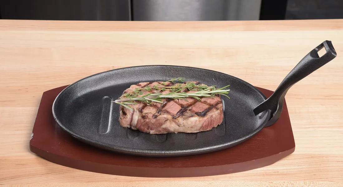 Cast iron skillet with a piece of seared steak topped with rosemary, resting on a wooden board