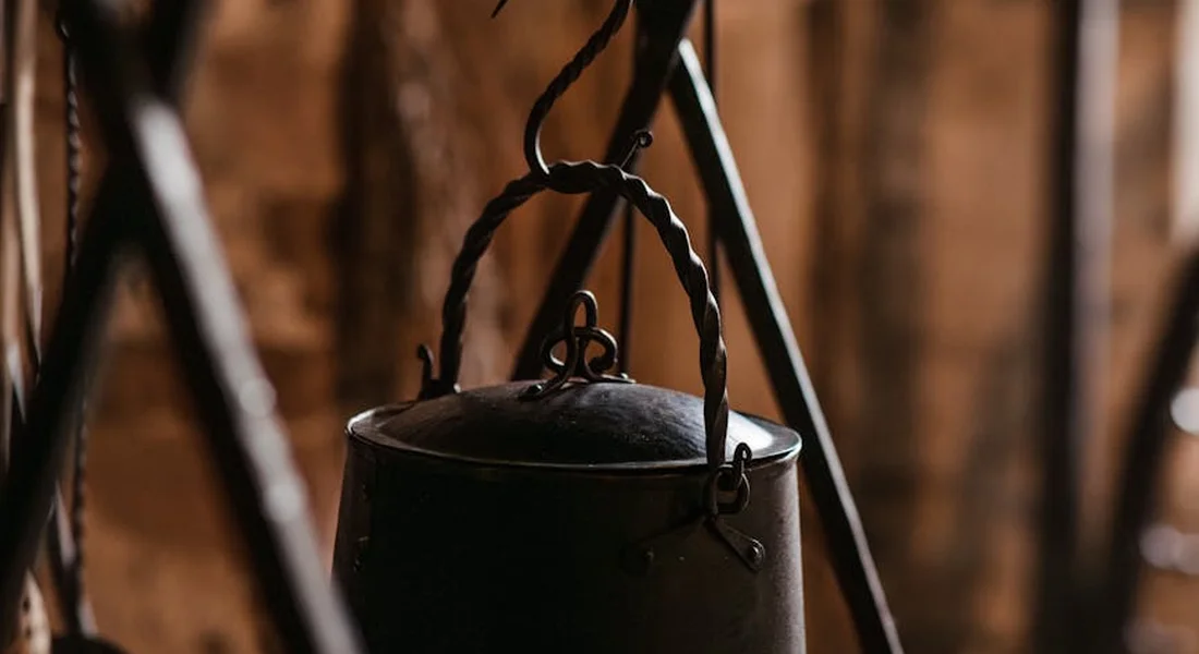 A dark metal pot with a curved handle hanging from a hook in a warm, rustic kitchen setting.