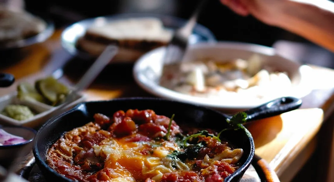 A cast-iron skillet with a tomato-based dish on a wooden board, surrounded by other plates on a table.