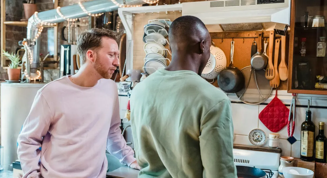 Two people standing at a kitchen counter discussing cooking, with pots, pans, and utensils hanging in the background.