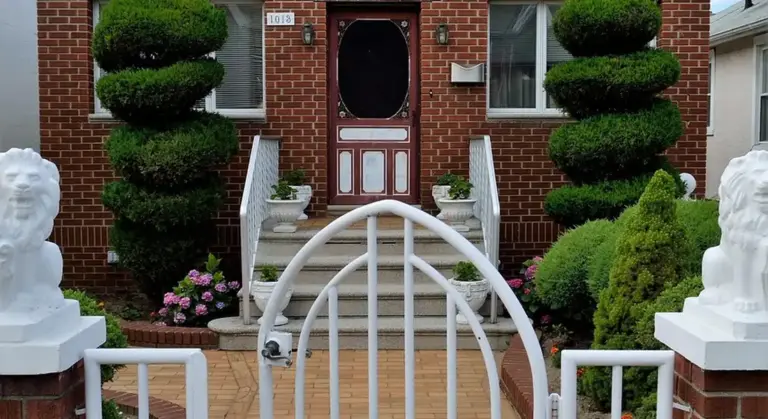 Front yard with a white wrought-iron gate, brick house, and manicured topiary shrubs flanking the entrance, with stone statues on either side.