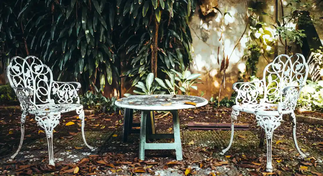 White wrought-iron outdoor chairs and a round table in a lush garden, with fallen leaves on the ground.