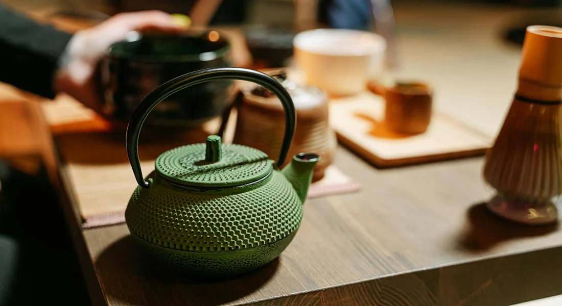 A textured green cast-iron teapot sits on a wooden table with blurred kitchen items in the background.