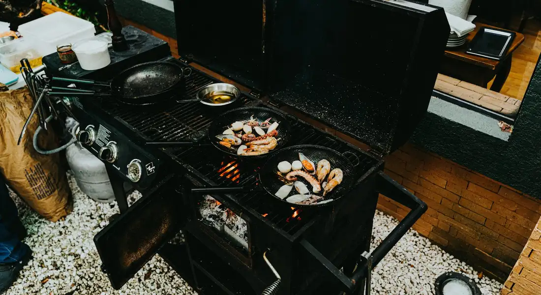 Top-down view of an outdoor charcoal grill with two cast-iron skillets on the grates, cooking food.