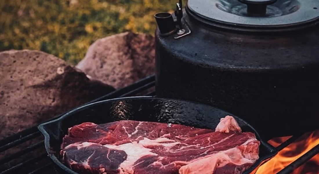 Raw beef steaks in a cast-iron skillet on a barbecue grill with a kettle in the background.