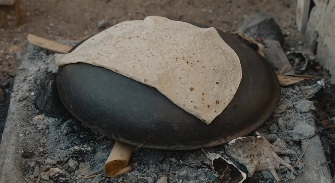 A weathered Griswold cast iron lid sits in a dirt and ash-filled outdoor area, with a light tan sheet draped over its surface.