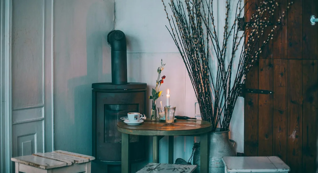 Cozy kitchen corner with a wood-burning stove, a round wooden table set with cups, and tall branches in a vase.