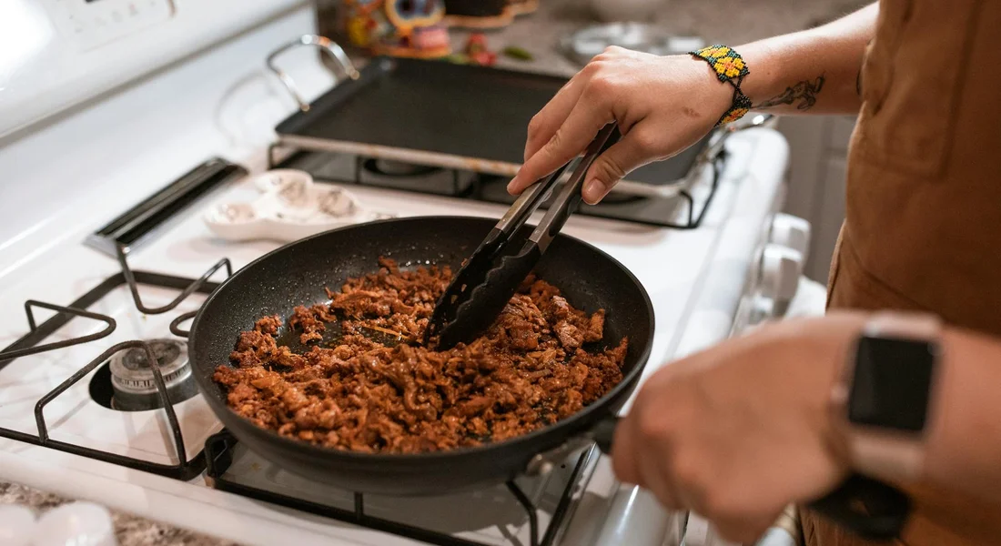 Person cooking in a kitchen using a cast-iron skillet on a gas stove; hands holding tongs, visible tattoo on forearm and a smartwatch on the other wrist