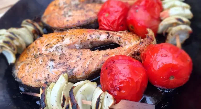 Close-up of grilled food on a cast-iron grill, featuring a seasoned piece of meat, onions, and bright red tomatoes.