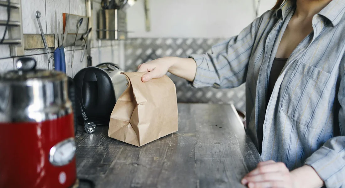 A person wearing a striped shirt reaches toward a brown paper bag on a kitchen counter next to a red kettle and a coffee maker, with a metal backsplash.