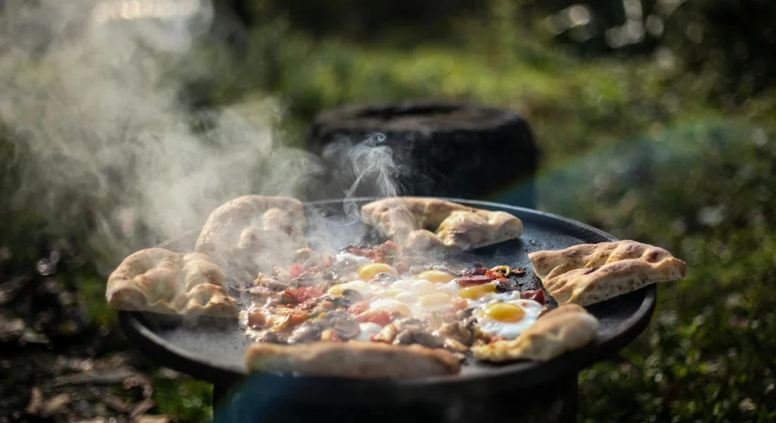 Cast iron skillet outdoors on a heat source, with bread rounds around the edge and eggs in the center, steam rising from the hot food.