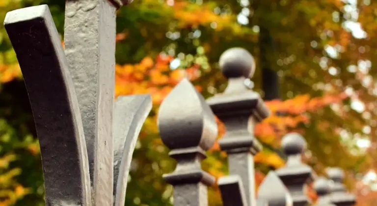 Close-up of wrought iron fence posts with decorative finials, blurred autumn leaves in the background.