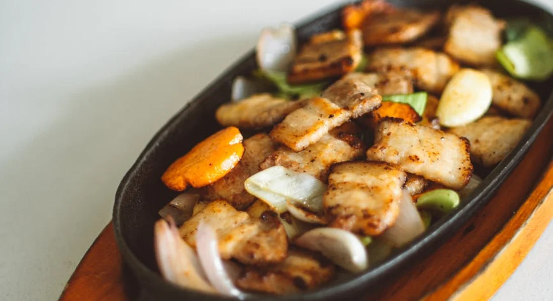 Cast iron skillet filled with seared meat and vegetables (onions and potatoes) cooking on a wooden surface.