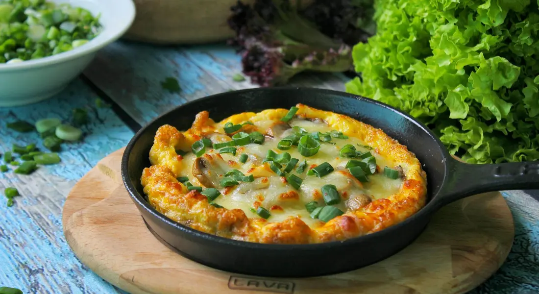 Cast iron skillet on a wooden board filled with a cheesy egg dish, with leafy greens in the background; a representative image of cookware used before or during seasoning.