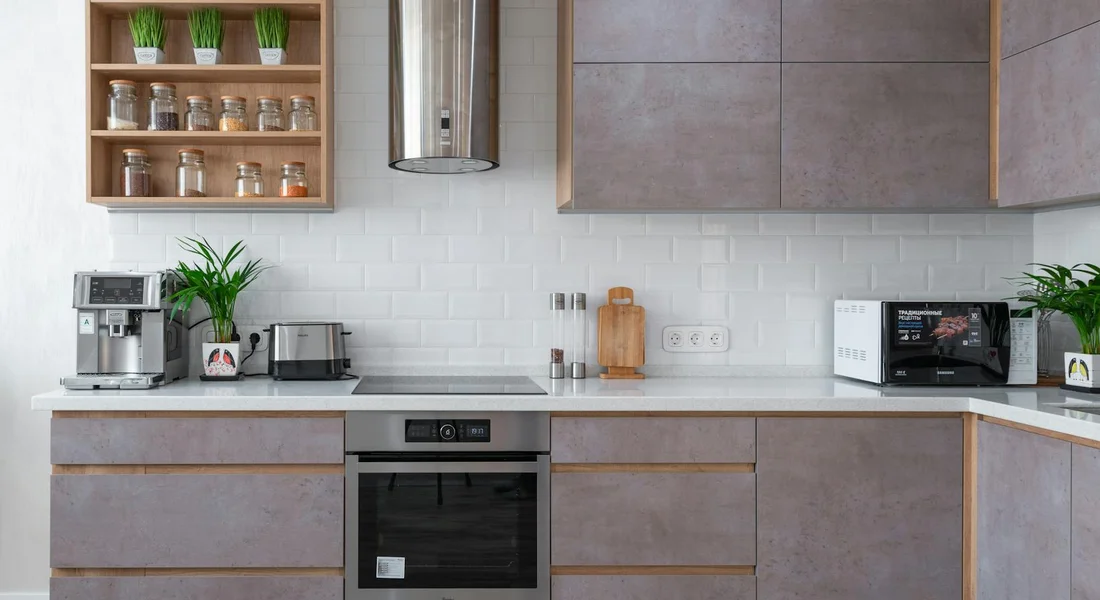 Modern kitchen with gray cabinetry, white subway tile backsplash, range hood, and countertop appliances including a coffee machine, toaster, microwave, and small potted plants.