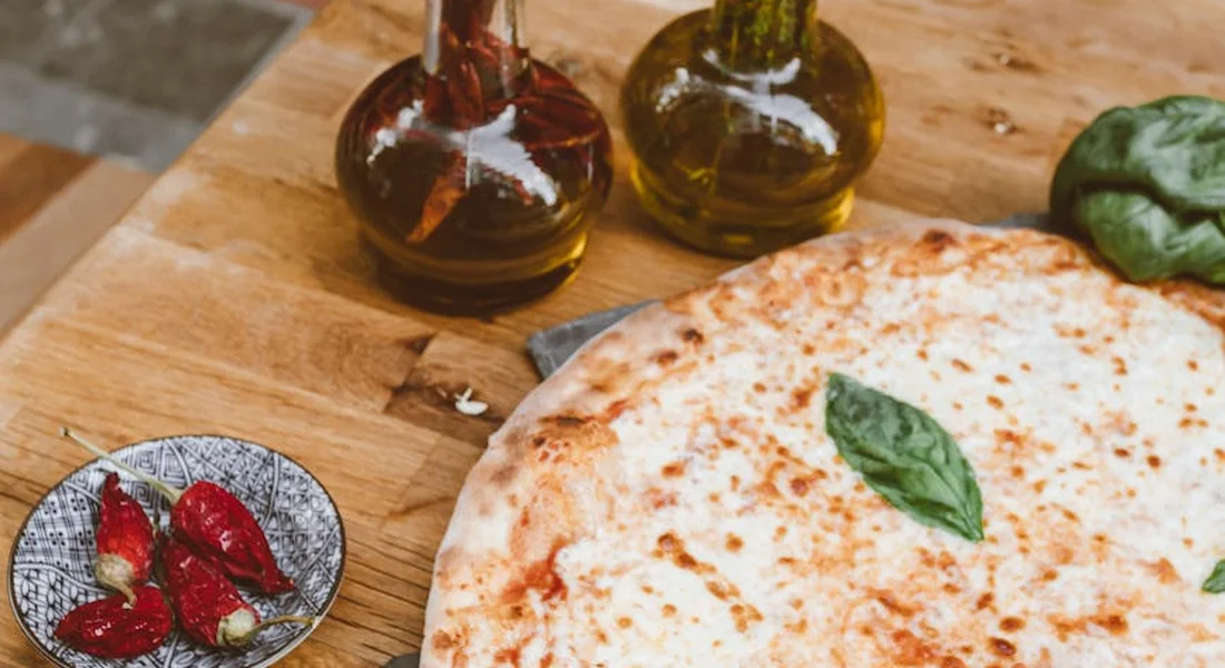 Amber glass oil bottles on a wooden table beside a pizza with a fresh basil leaf.