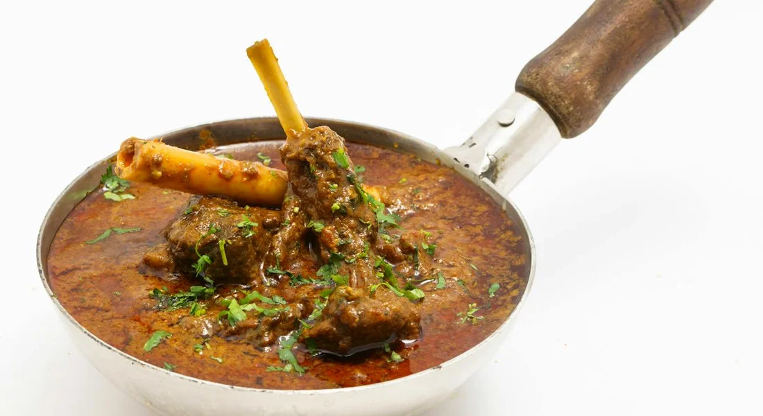 A shallow metal skillet with red braised sauce and chunks of meat, garnished with herbs, featuring a wooden-handled handle.