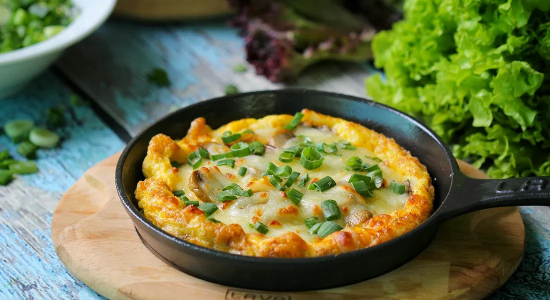 Cast iron skillet with bubbling pancake or omelette topped with chopped scallions on a wooden board.