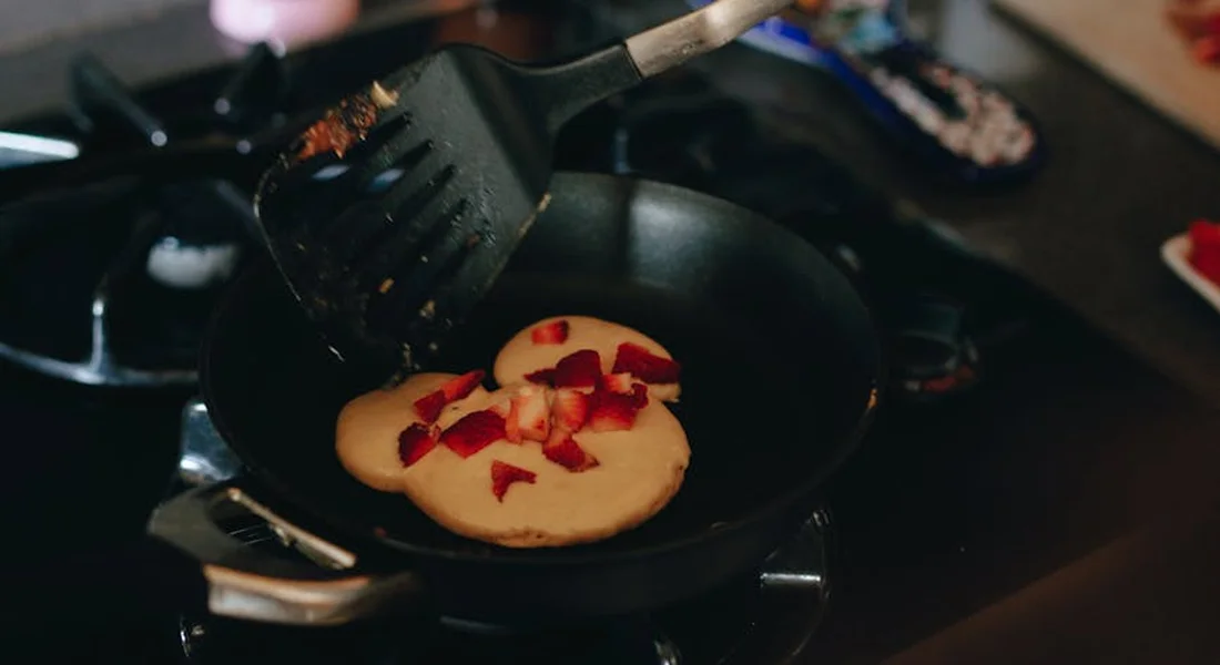 A pancake cooking in a cast-iron skillet with a spatula hovering above and bits of fruit on top.
