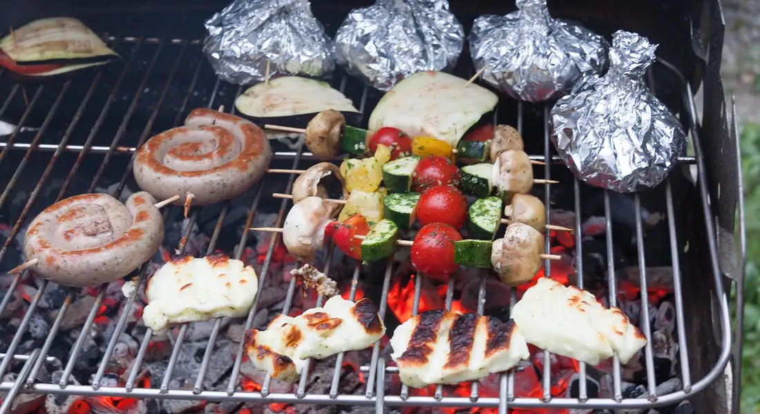 Grill with skewered vegetables and sausages cooking on a porcelain-enameled cast iron grill, with foil packets in the background.