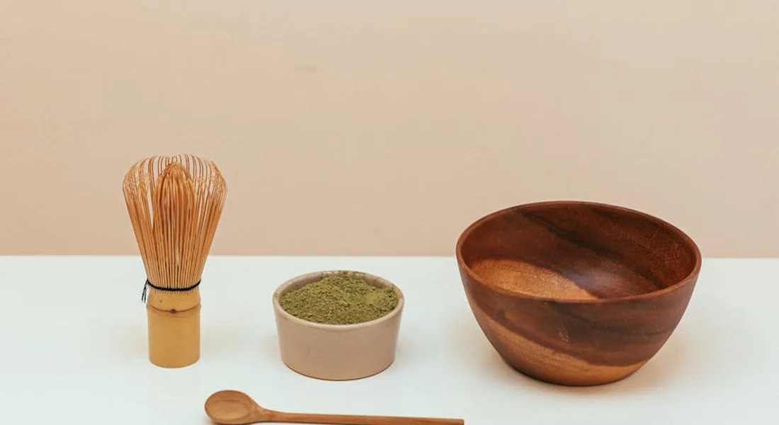 A wooden mixing bowl, a wooden spoon, a small bowl of green powder, and a bamboo whisk arranged on a light countertop, illustrating utensils used with cast iron cookware.