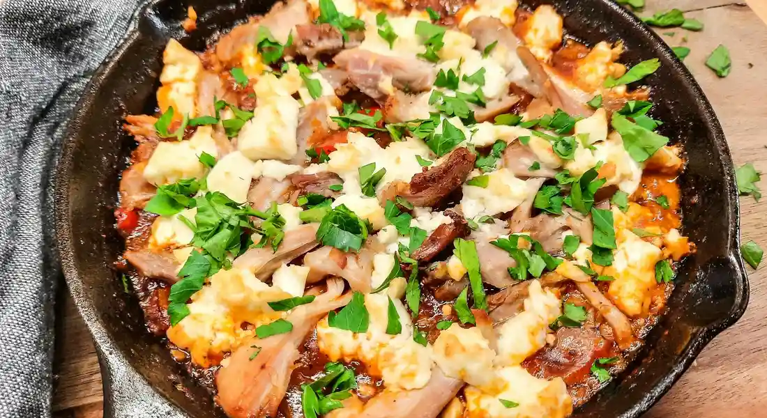 Cast-iron skillet containing a cheesy, meat-and-herb dish, topped with chopped parsley, on a wooden surface.