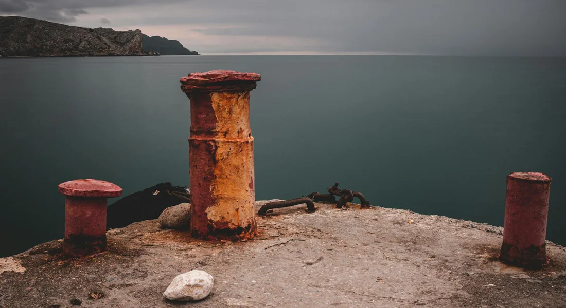 Three rusted cast iron bollards on a weathered concrete pier with calm water in the background.