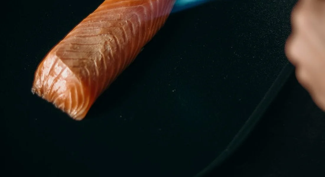 Close-up of a salmon fillet being sliced on a dark surface with a knife; a blurred hand is visible in the background.
