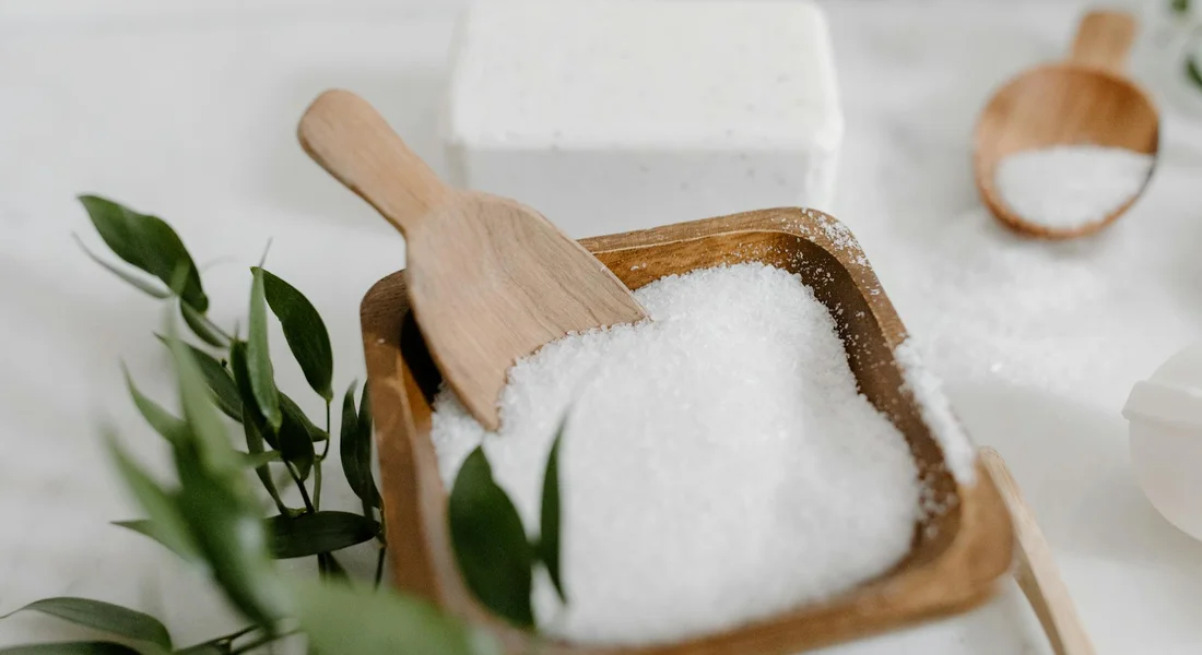 Coarse salt in a wooden scoop on a light kitchen surface with green leaves nearby