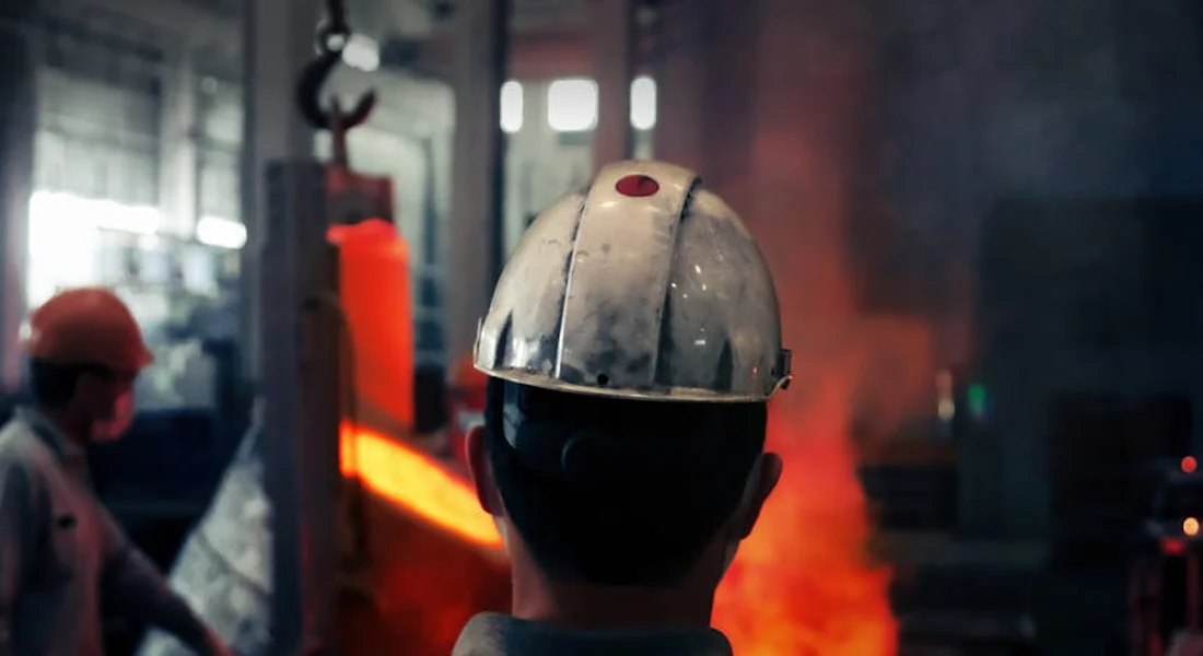 Worker wearing a hard hat in a workshop, watching a glowing metal surface being cleaned by a sandblaster.