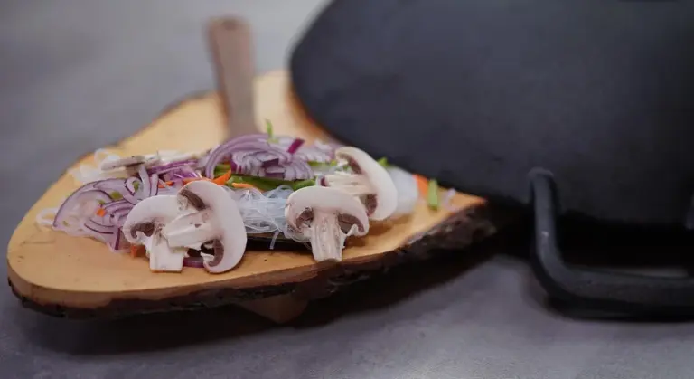 Wooden board with sliced red onions, mushrooms, and garlic next to a black cast iron lid.