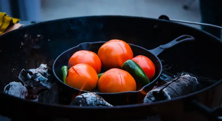 Cast iron skillet on a charcoal grill containing bright orange tomatoes and a green pepper.
