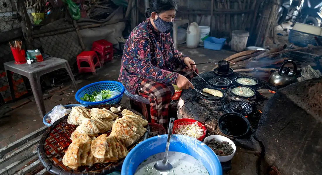 A masked cook in a rustic outdoor kitchen tends a row of pans and a blue bowl of ingredients, with flatbreads on a grill and various sauces nearby.