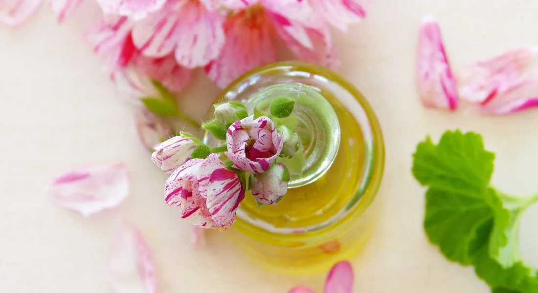 Close-up of a small glass bottle filled with oil, topped with a pink flower, with pink petals and a green leaf on a light surface.