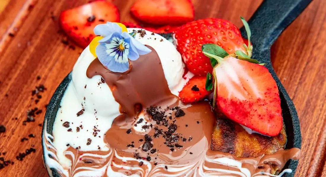 Dessert in a skillet with vanilla ice cream, melted chocolate, fresh strawberry slices, and a blue edible flower on a wooden surface.